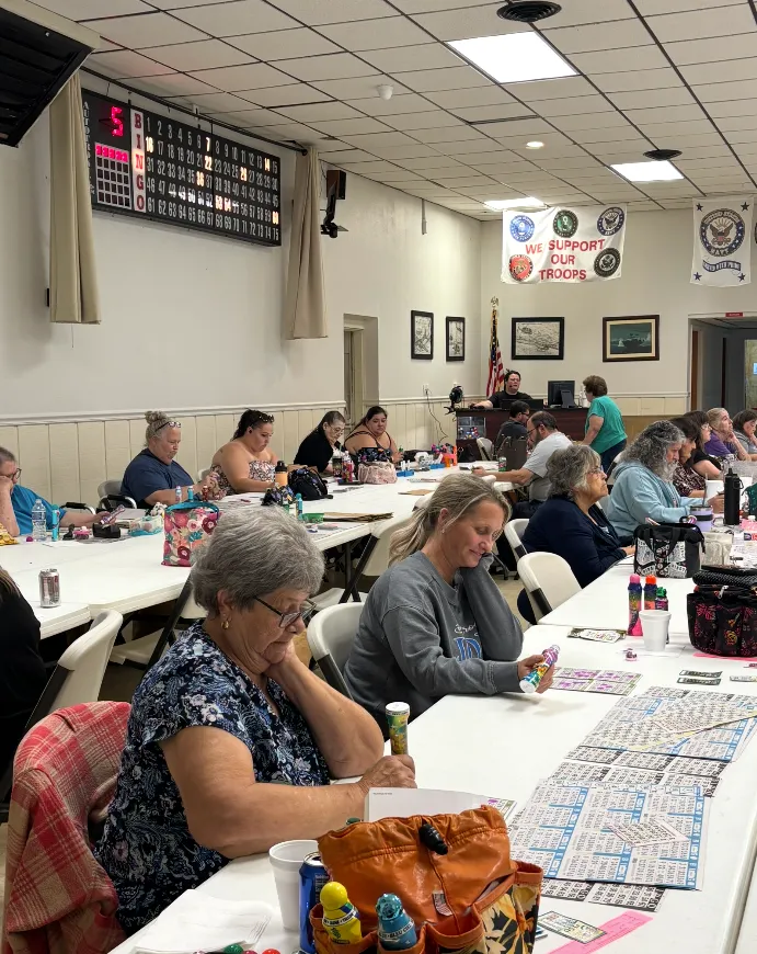 Players filling out bingo cards at a Thursday night session at Post 4093, with the call board and a We Support Our Troops banner in the background